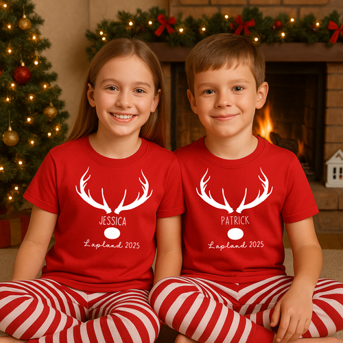 Two children wearing red shirts with white antler designs and names, sitting in front of a decorated Christmas tree and fireplace. Cosy Christmas Lapland pyjamas – personalised family Lapland 2025 PJs set, ideal as a Lapland trip reveal outfit, matching Lapland holiday outfits, or a unique personalised Lapland gift idea for travel.