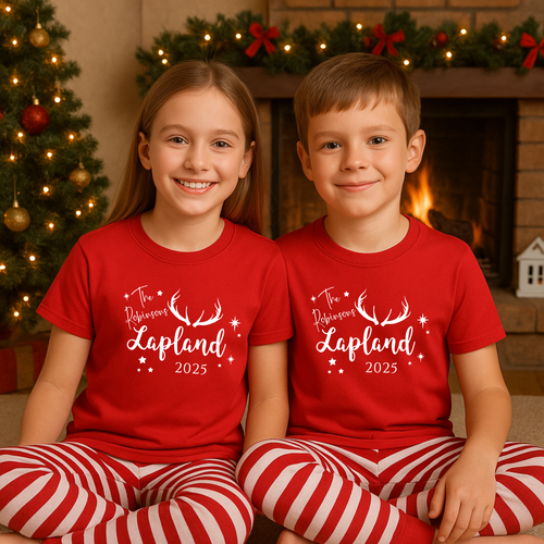 Two children wearing red shirts with Lapland 2025 Text, sitting in front of a decorated Christmas tree and fireplace. Cosy Christmas Lapland pajamas for matching family Lapland pyjamas