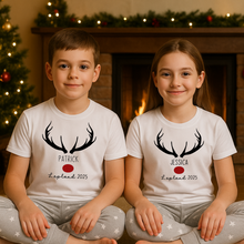 Load image into Gallery viewer, Two children wearing matching shirts with antlers and names in front of a fireplace and Christmas tree.
Personalised Lapland holiday PJs for families – cosy Christmas Lapland pyjamas that double as a Lapland trip reveal outfit, matching Lapland holiday outfits, and a thoughtful personalised Lapland gift idea.