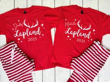 Load image into Gallery viewer, Two red t-shirts with &#39;Lapland 2025&#39; text and matching red and white striped pajama sets on a wooden surface. Cosy Christmas Lapland pajamas for matching family Lapland pyjamas