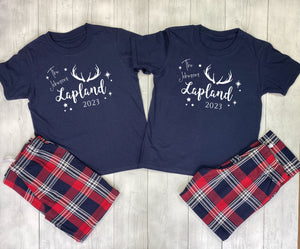 Two navy blue t-shirts with 'The Adventure is in Lapland 2023' text and plaid shorts on a wooden surface.