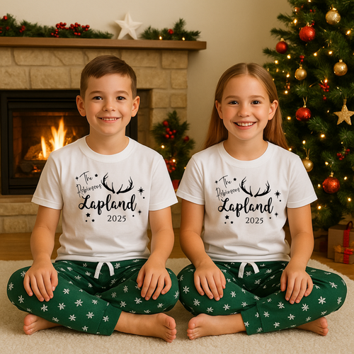 Personalised Lapland holiday PJs for Lapland trip reveal matching outfits.Two children wearing matching pajamas with 'Lapland 2025' text in front of a Christmas tree and fireplace.