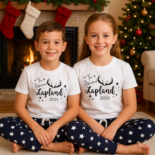 Two children wearing matching shirts personalised with with their family name and 'Lapland 2025' text, sitting in front of a fireplace and Christmas tree. Matching family Lapland pyjamas as Lapland holiday matching pyjamas sets