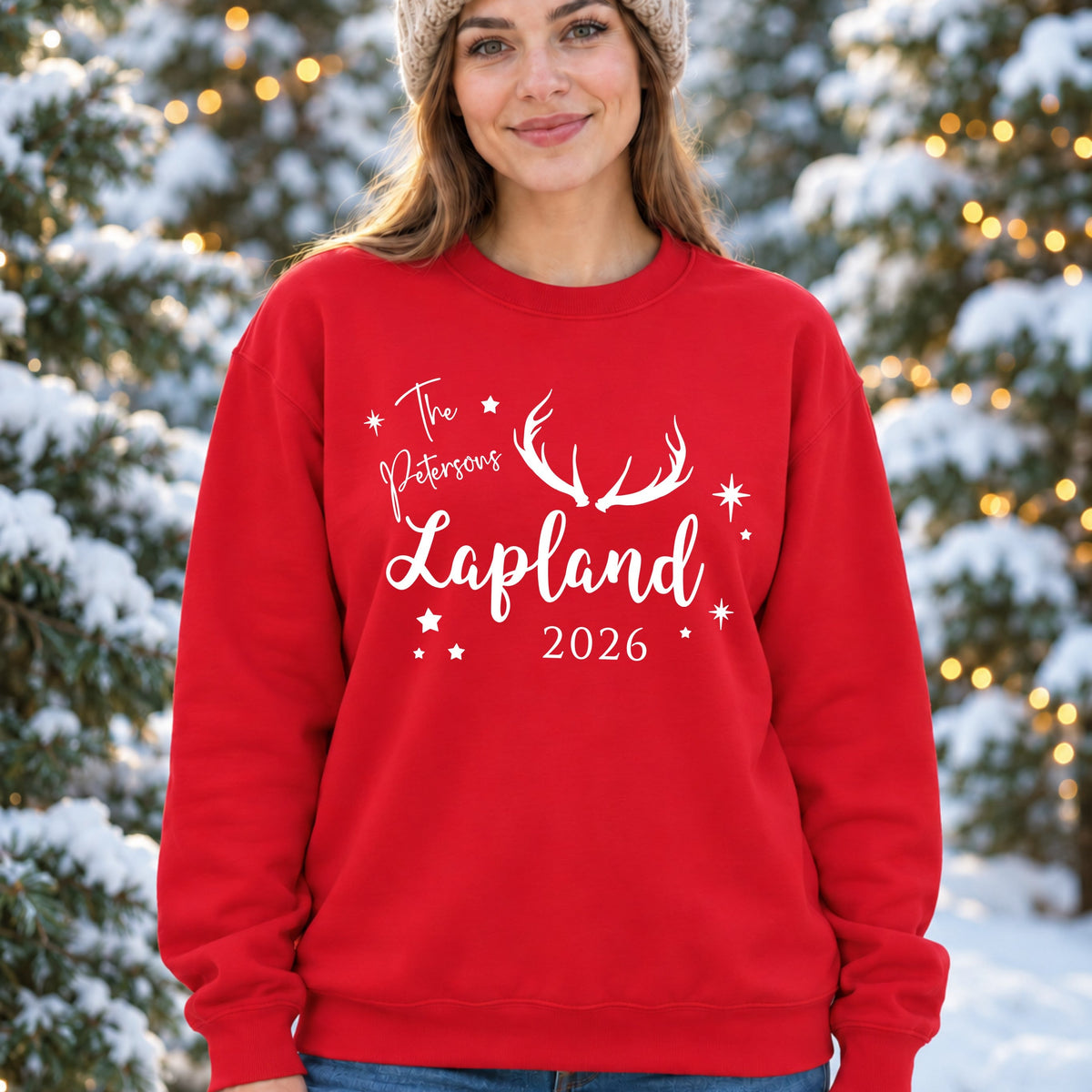 A woman wearing a red crewneck sweatshirt with a white graphic that reads 'The Pattersons Lapland 2026' with antler artwork, standing in a snowy outdoor setting with decorated evergreen trees.