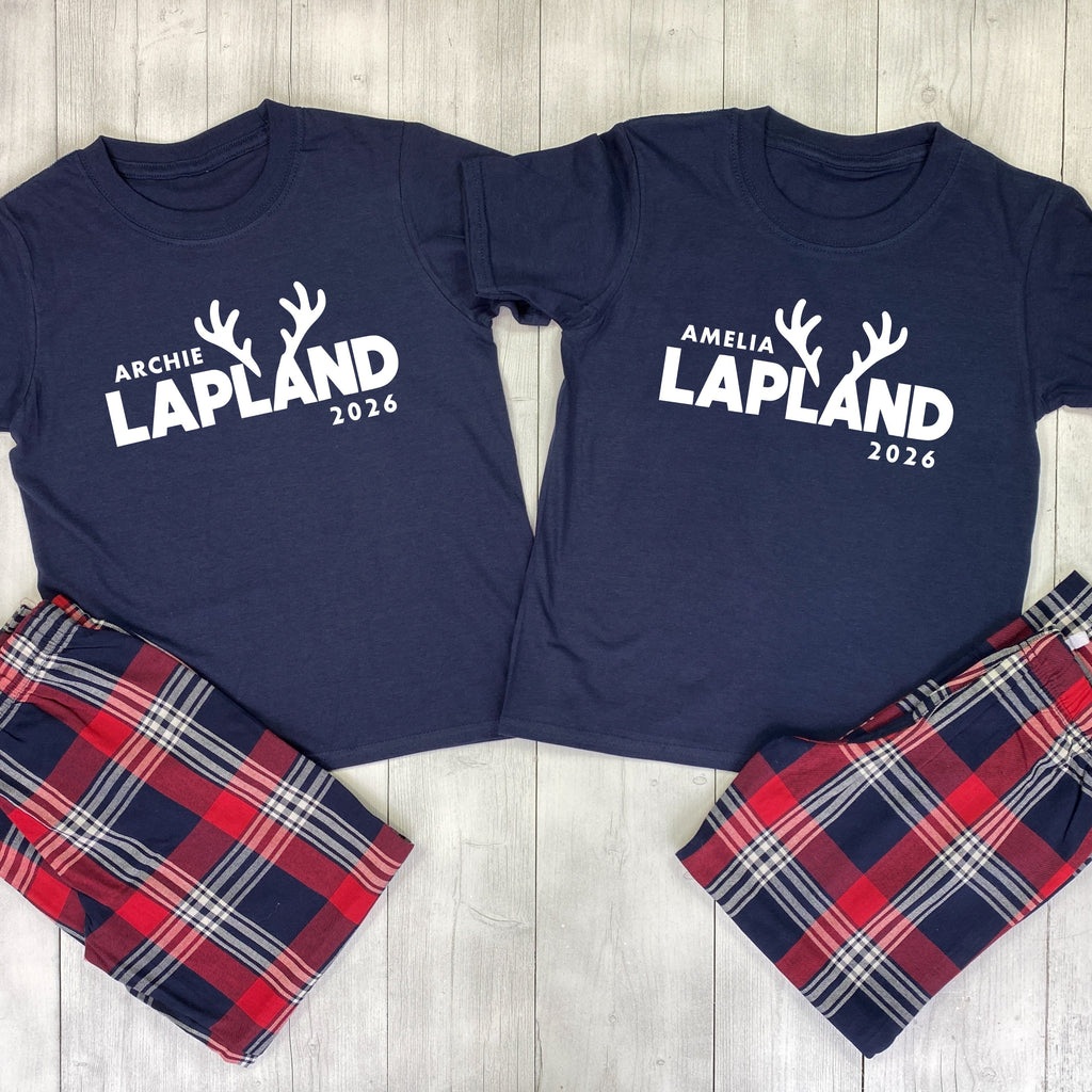 Two navy blue matching pajama shirts laid flat on a light wood background, each printed with white text and reindeer antlers reading names and '2026', paired with red, navy, white, and gray plaid pajama pants.