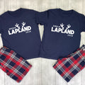 Two navy blue matching pajama shirts laid flat on a light wood background, each printed with white text and reindeer antlers reading names and '2026', paired with red, navy, white, and gray plaid pajama pants.