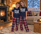 Two children standing in a cozy log cabin living room wearing matching navy blue t-shirts and red, navy, and white plaid pajama pants. The shirts feature a white reindeer graphic and the text 'SANTA LALAND'.