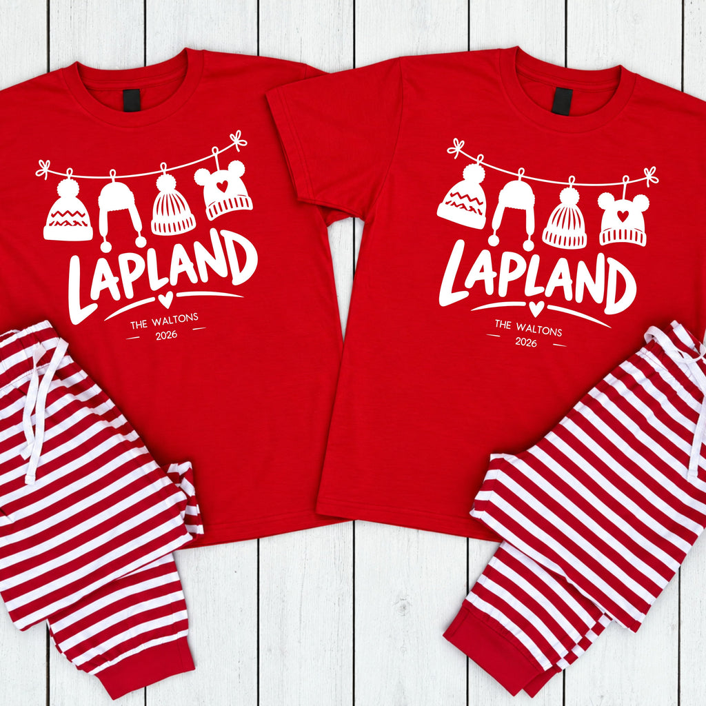 Two matching red pajama sets laid flat on a white wood plank background. Each set includes a short-sleeve shirt with a white graphic of knit hats hanging on a line above the text 'LAPLAND' and smaller text 'THE WALKERS 2026', paired with striped pants in red and white.