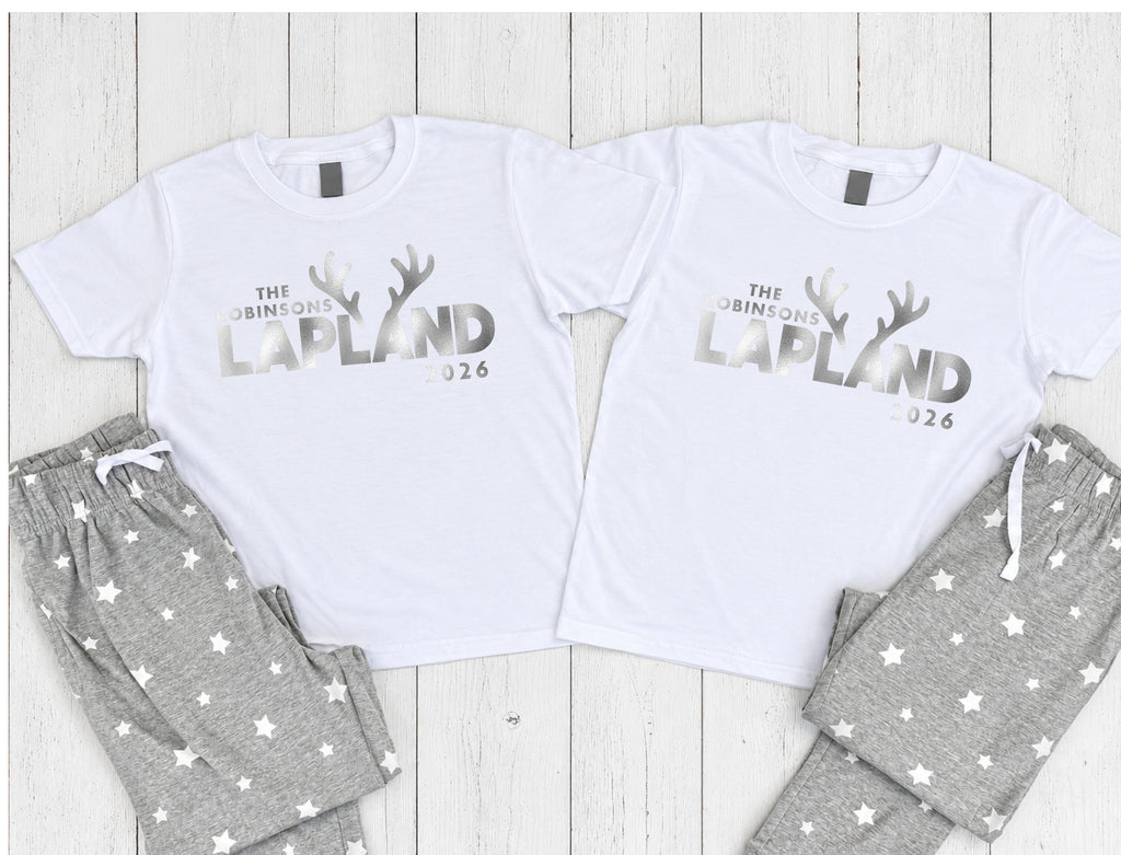 Two matching white short-sleeve pajama shirts with gray text reading 'THE GRIMFOLD LAPLAND 2026' and gray antler graphics, paired with gray star-print pajama pants, arranged on a white wood plank background.