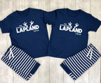 A matching couple's sleepwear set laid flat on a light wood background. The set includes two navy blue short-sleeve t-shirts with white text reading 'THE ROBINSONS LAPLAND 2026' and antler graphics, paired with matching navy and white striped shorts.