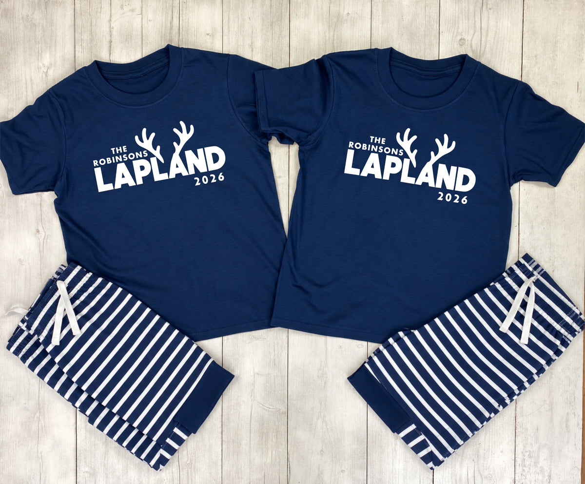 A matching couple's sleepwear set laid flat on a light wood background. The set includes two navy blue short-sleeve t-shirts with white text reading 'THE ROBINSONS LAPLAND 2026' and antler graphics, paired with matching navy and white striped shorts.