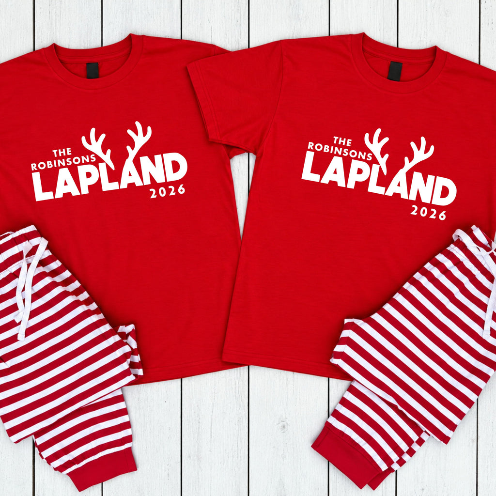 Two matching red short-sleeve pajama shirts with white text reading 'THE ROBINSONS LAPLAND 2026' and reindeer antler graphics, paired with red-and-white striped pajama pants, laid flat on a white wood plank background.