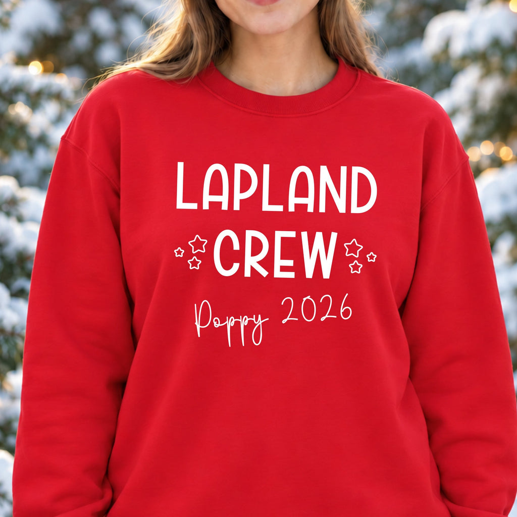 Woman wearing a red Lapland Crew sweatshirt with white lettering and outlined stars, personalised with Poppy and 2026, in snowy woodland.