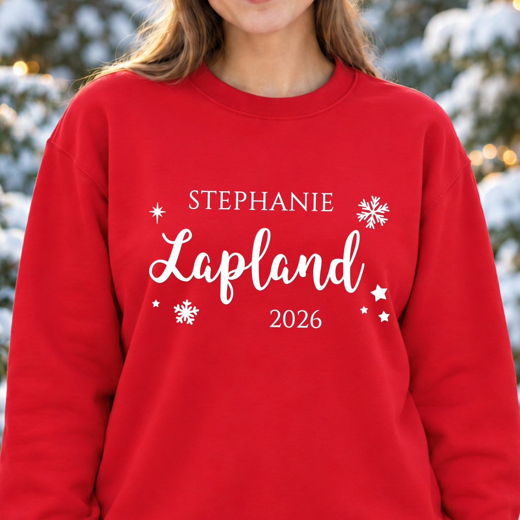 Woman wearing a red personalised Lapland 2026 sweatshirt with white snowflakes, stars and the name Stephanie in a snowy woodland with fairy lights.