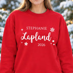 Woman wearing a red personalised Lapland 2026 sweatshirt with white snowflakes, stars and the name Stephanie in a snowy woodland with fairy lights.