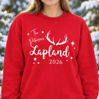 A woman wearing a red crewneck sweatshirt with white text reading 'The Petersons Lapland 2026' and white antler graphics, paired with a beige knit beanie and blue jeans, standing in a snowy outdoor setting with blurred Christmas trees and warm lights.