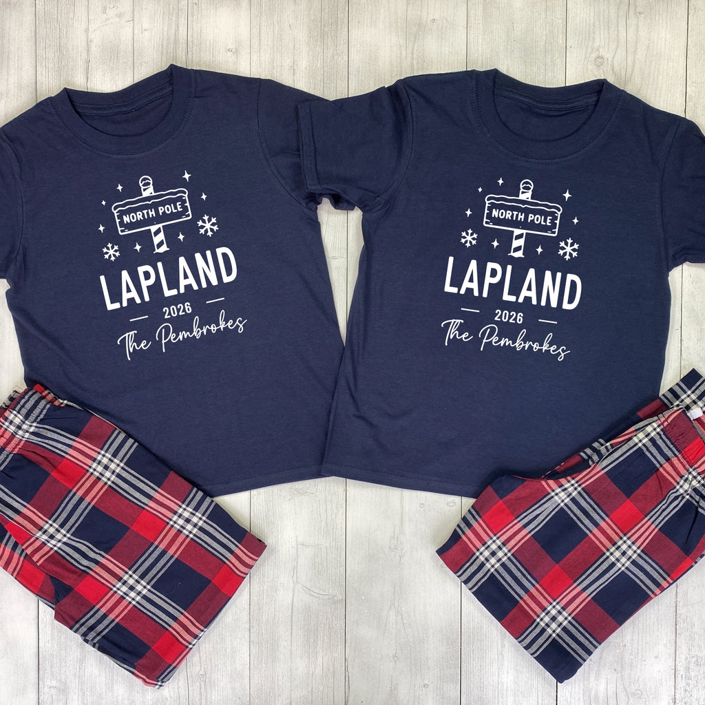 Two navy blue matching t-shirts with white graphic prints reading 'NORTH POLE 1 LAPLAND 2026 The Pembrokes' are laid flat on a light wood background, paired with red, navy, and white plaid shorts.