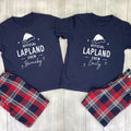 A matching couple's pajama set laid flat on a light wood background, featuring navy blue short-sleeve shirts with white snowman graphics and names 'KAREN' and 'JAMES', paired with red, navy, and white plaid shorts.