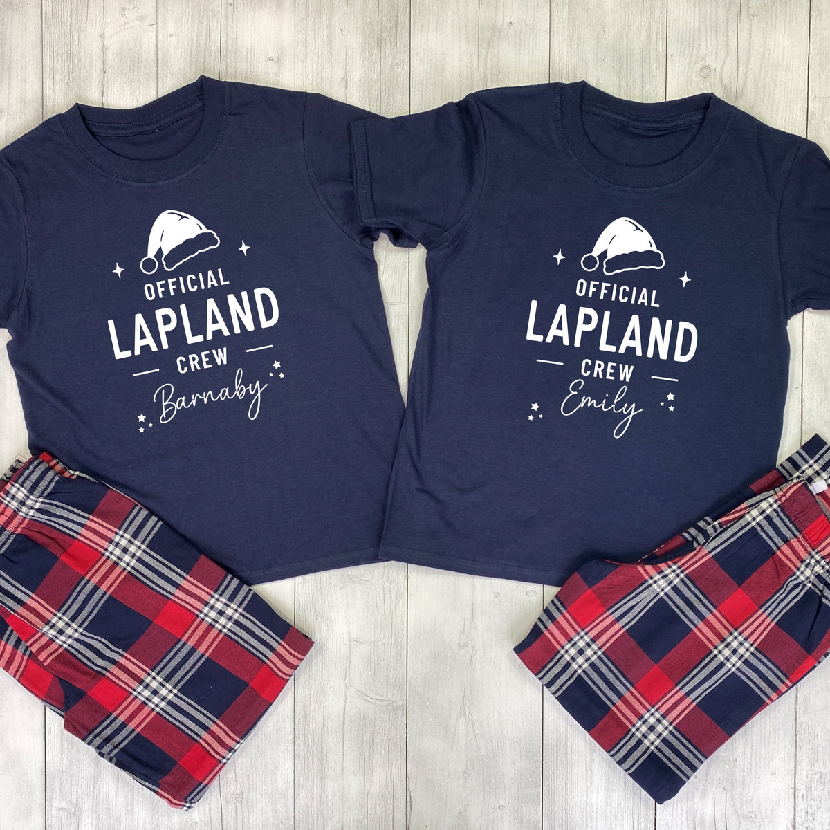 A matching couple's pajama set laid flat on a light wood background, featuring navy blue short-sleeve shirts with white snowman graphics and names 'KAREN' and 'JAMES', paired with red, navy, and white plaid shorts.