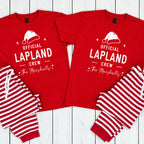 Two matching red pajama sets laid flat on a white wood background. Each set includes a short-sleeve crew-neck shirt with white text reading 'OFFICIAL LAPLAND CREW' and 'The Marshalls' beneath it, plus a white Santa hat graphic, and striped pants in red and white.