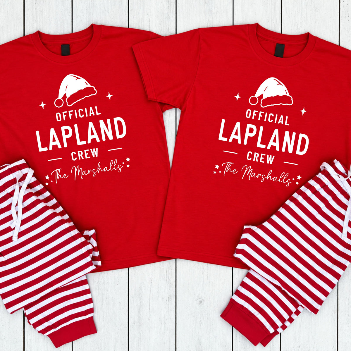 Two matching red pajama sets laid flat on a white wood background. Each set includes a short-sleeve crew-neck shirt with white text reading 'OFFICIAL LAPLAND CREW' and 'The Marshalls' beneath it, plus a white Santa hat graphic, and striped pants in red and white.