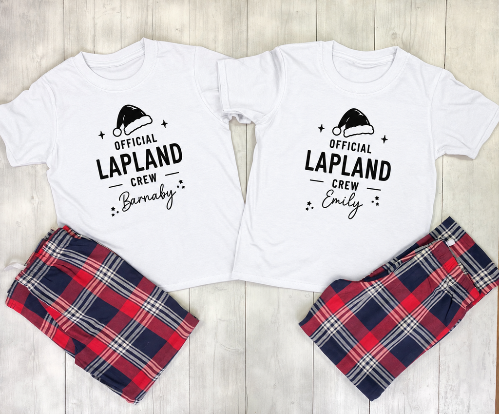 Two white graphic pajama shirts laid flat on a light wood background, each paired with plaid pajama pants. The left shirt is white with black text reading 'Official Lapland Crew Barnaby' and a Santa hat graphic; the right shirt is white with black text reading 'Official Lapland Crew Emily' and a Santa hat graphic.