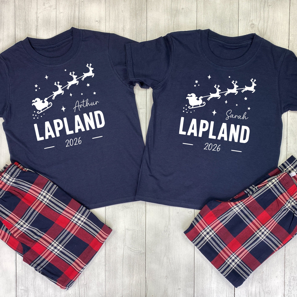 A matching Christmas-themed pajama set laid flat on a light wood background, featuring two navy blue short-sleeve shirts with white holiday graphics and names, paired with red, navy, and white plaid pants.