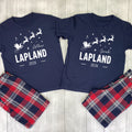 A matching Christmas-themed pajama set laid flat on a light wood background, featuring two navy blue short-sleeve shirts with white holiday graphics and names, paired with red, navy, and white plaid pants.