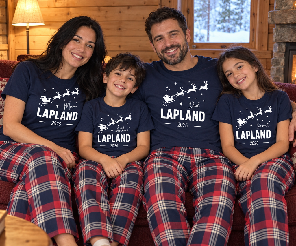 A family of four wearing matching navy blue t-shirts and red, navy, and white plaid pajama pants, sitting together on a couch in a cozy wood-paneled room. The shirts feature white text and graphics reading 'Mrs.