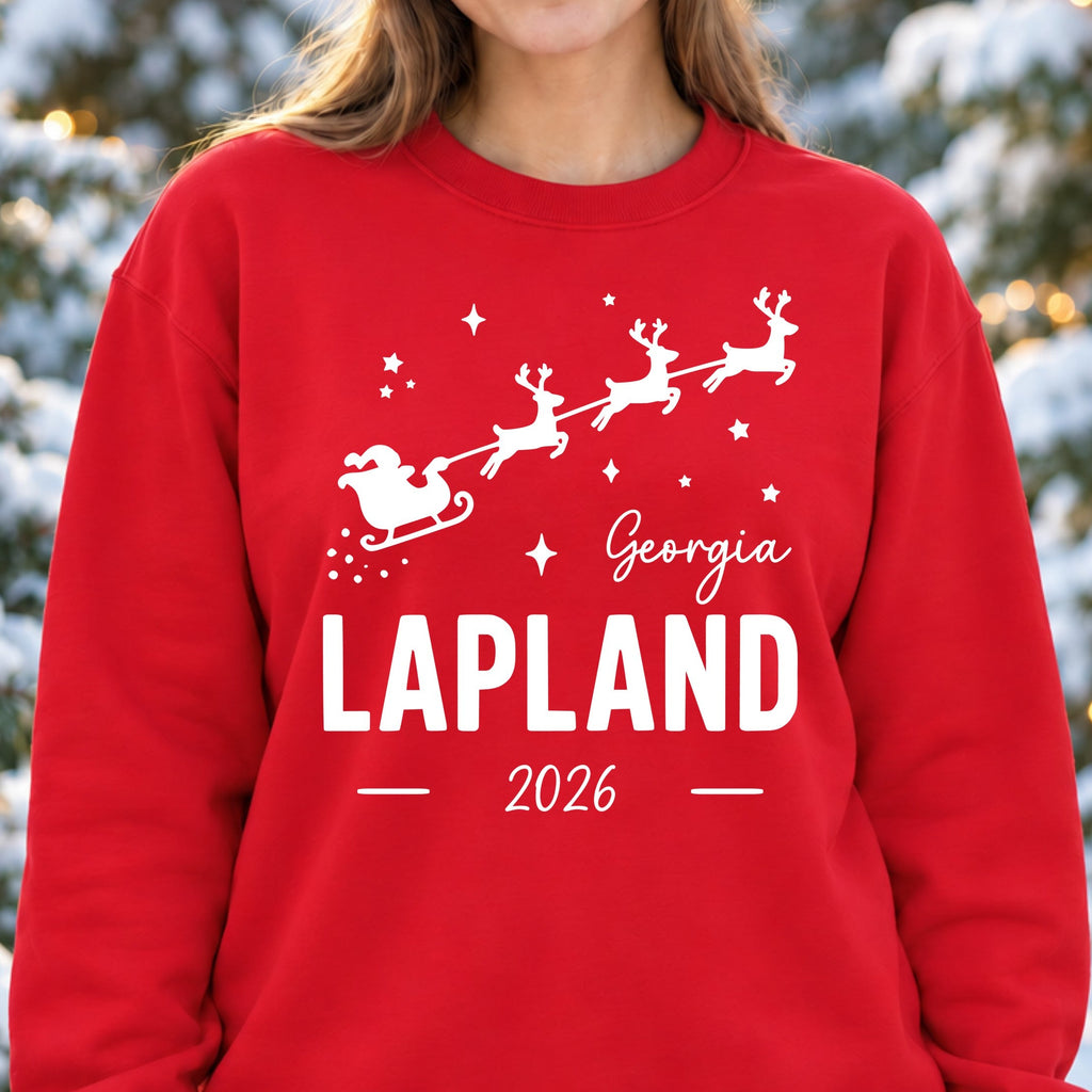 Woman wearing a red personalised Lapland 2026 sweatshirt with white flying Santa sleigh, stars and the name Georgia in snowy woodland.