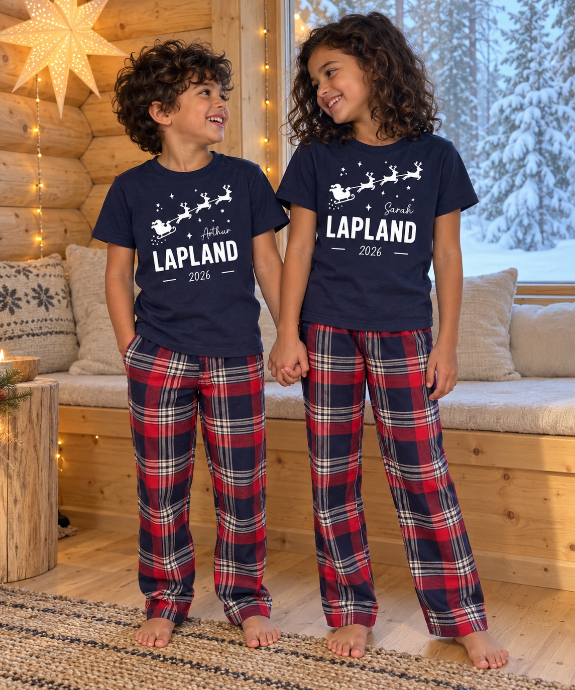 Two children standing indoors wearing matching navy holiday pajama sets with white Santa and reindeer graphics and red/navy plaid pants in a cozy log-cabin-style room.