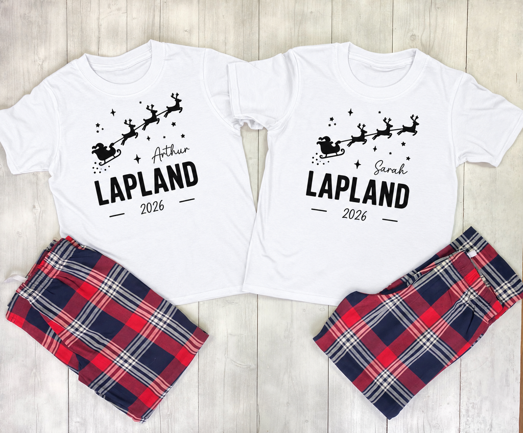 Two matching Christmas-themed pajama sets laid flat on a light wood background. Each set includes a white short-sleeve T-shirt with a black Santa and reindeer graphic and the name 'LAPLAND' plus 2026, paired with plaid shorts in red, navy, white, and gray.
