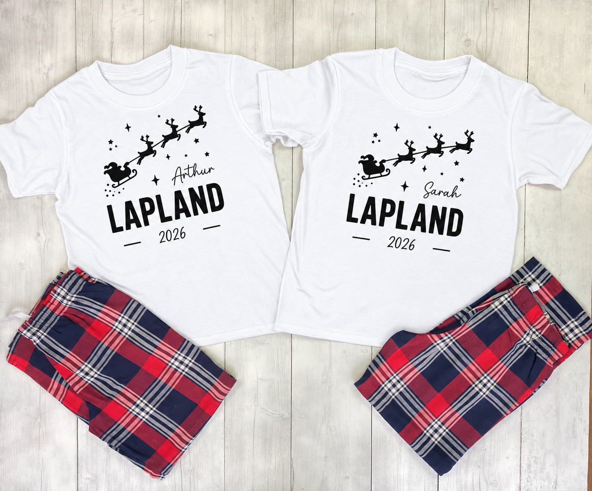 Two matching Christmas-themed pajama sets laid flat on a light wood background. Each set includes a white short-sleeve T-shirt with a black Santa and reindeer graphic and the name 'LAPLAND' plus 2026, paired with plaid shorts in red, navy, white, and gray.