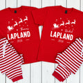 Two matching Christmas pajama sets laid flat on a white wood background. Each set includes a red short-sleeve T-shirt with a white holiday graphic and personalized text ('Thomas' and 'Rachel', 'LAPLAND', '2026') and red-and-white striped pants.